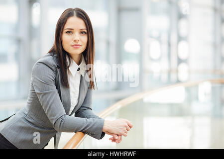 Portrait of beautiful brunette businesswoman looking at camera leaning on glass railing in modern business center Stock Photo