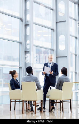 Group of  business people sitting in circle during meeting, man standing up giving presentation in glass hall of modern office building Stock Photo