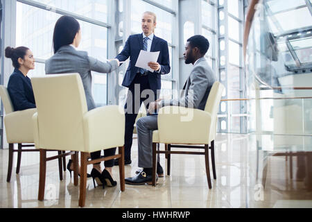 Group of  business people sitting in circle during meeting, man standing up and handing documents to woman partner after giving presentation in glass  Stock Photo