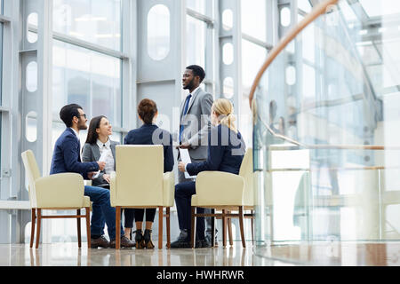 Group of  business people sitting in circle during meeting, African  man standing up giving speech  in glass hall of modern office building Stock Photo