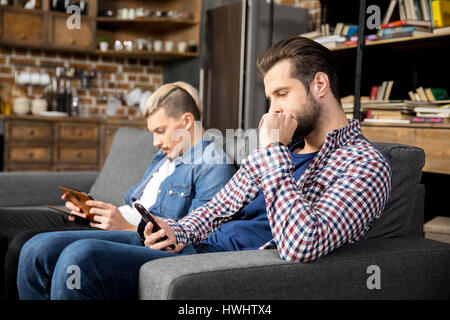 Two young men sitting on couch and using digital devices Stock Photo