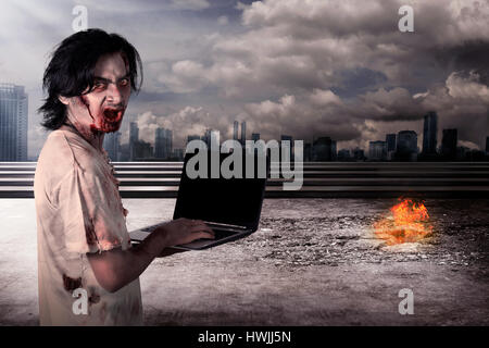 Creepy male zombie typing with laptop with city on fire background Stock Photo