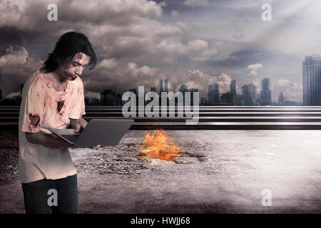 Creepy male zombie typing with laptop with city on fire background Stock Photo