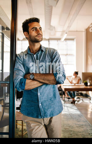 Portrait of young businessman standing at door while colleagues working in background. Young male at startup office. Stock Photo