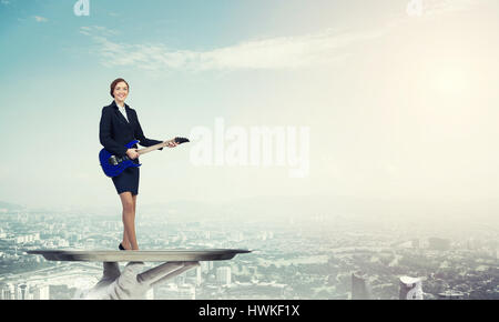 Attractive businesswoman on metal tray playing electric guitar against cityscape background Stock Photo