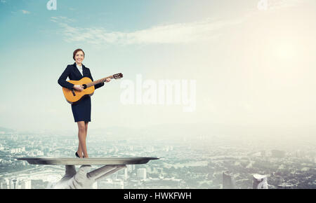 Attractive businesswoman on metal tray playing acoustic guitar against cityscape background Stock Photo