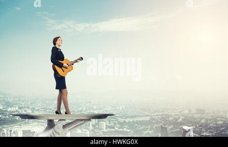 Attractive businesswoman on metal tray playing acoustic guitar against cityscape background Stock Photo