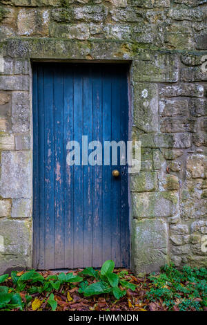 Faded and peeling old blue painted wooden door with brass door knob against lichen covered stone wall Stock Photo