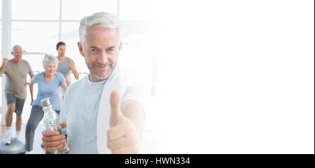 Man showing thumbs up sign with people exercising in background at fitness studio Stock Photo