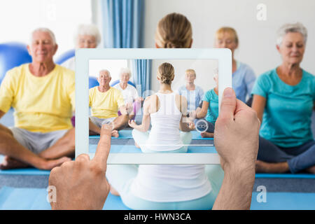 Hand holding tablet pc against instructor performing yoga with seniors Stock Photo