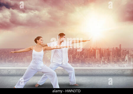 Peaceful couple in white doing yoga together in warrior position against balcony overlooking city Stock Photo