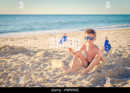 Cute smiling boy sitting on the beach and holding Australian flags on Australia day Stock Photo