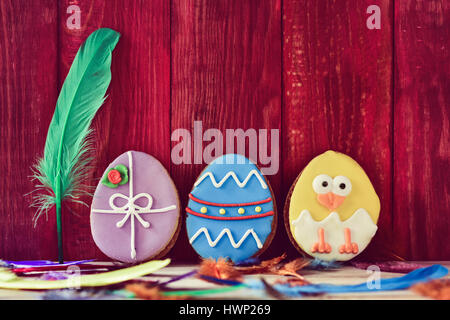 a pile of different some cookies patterned as different decorated easter eggs against a red wooden background, and feathers of different colors sprink Stock Photo