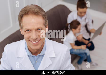 At my workplace. Handsome smiling distinguished doctor standing in his office and looking right in the camera while his colleague consulting little gi Stock Photo