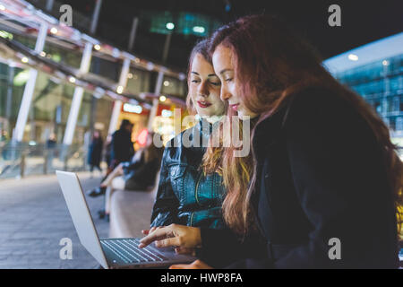 Two young beautiful caucasian women friends outdoor in the city night using computer, tapping keyboard - business, technology, social network concept Stock Photo