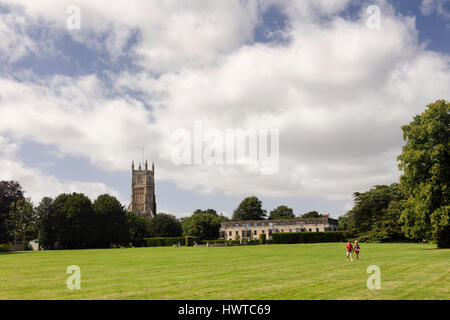 Parish Church of John the Baptist in Cirencester from Cirencester Abbey Grounds. Two people are walking across a park in the foreground. Stock Photo