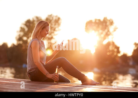 Alone lady sitting on dock near water with bottle and cellphone at sunset Stock Photo