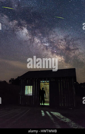 A man is silhouetted against a back lit lonely shed under a dramatic night sky with the milky way above. Stock Photo