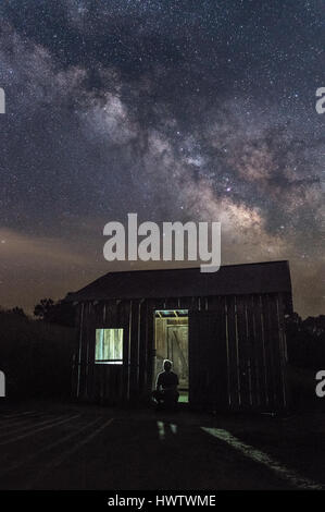 A man is silhouetted against a back lit lonely shed under a dramatic night sky with the milky way above. Stock Photo