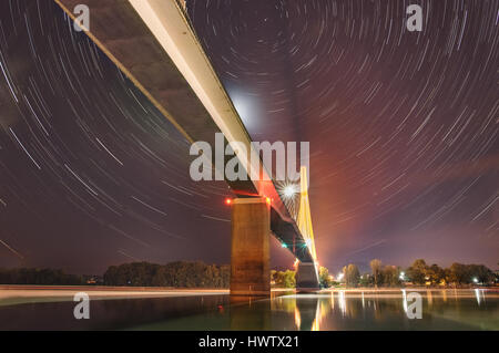 The rotation of the Earth produce star trails over long exposures at night above the large cable-stay bridge shrouded in light pollution. Stock Photo