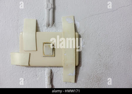 Dirty broken electrical socket with tapes on usable light switch and the small long crack white wall as a background Stock Photo