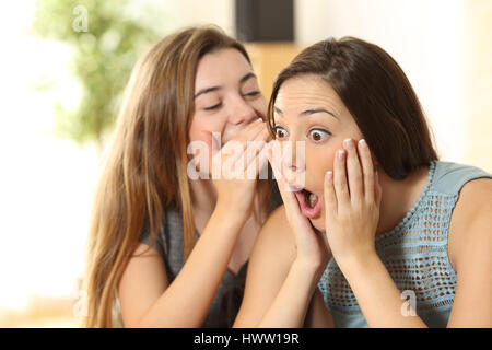 Portrait of a girl telling secrets to her amazed friend sitting on a couch in the living room at home Stock Photo