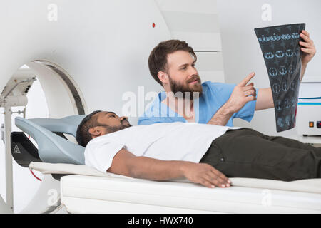 Young male radiologist showing X-ray to patient lying on CT scanner at hospital Stock Photo