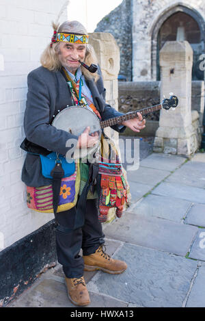 An quirky street entertainer wearing a headdress and smoking a pipe while playing his banjo in Canterbury, Kent, UK. Stock Photo