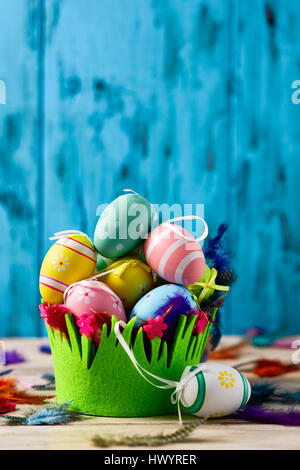 a pile of different decorated easter eggs in a basket decorated as grass and feathers of different colors sprinkled on a rustic surface, against a blu Stock Photo
