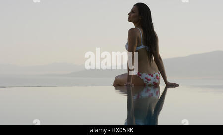 Rear view of woman in bikini sitting at edge of infinity pool overlooking horizon and remote landscape Stock Photo