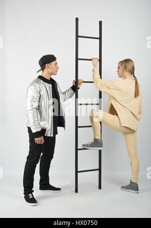 Modern fashionable hipster couple with ladder. Young muscular man wearing black clothes and sneakers with girl in yellow clothes on white background.  Stock Photo