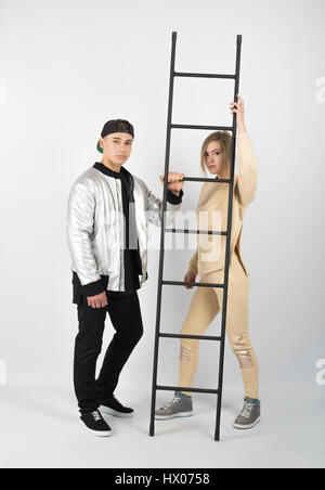 Modern fashionable hipster couple with ladder. Young muscular man wearing black clothes and sneakers with girl in yellow clothes on white background Stock Photo