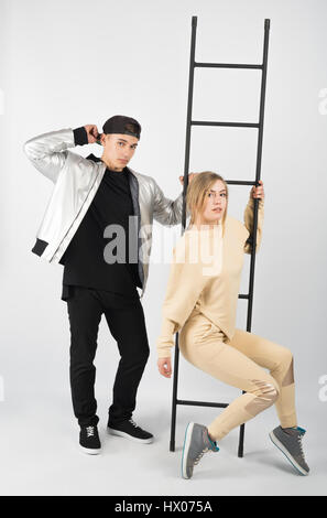 Modern fashionable hipster couple with ladder. Young muscular man wearing black clothes and sneakers with girl in yellow clothes on white background.  Stock Photo