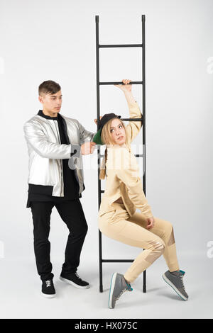 Modern fashionable hipster couple with ladder. Young muscular man wearing black clothes and sneakers with girl in yellow clothes on white background.  Stock Photo