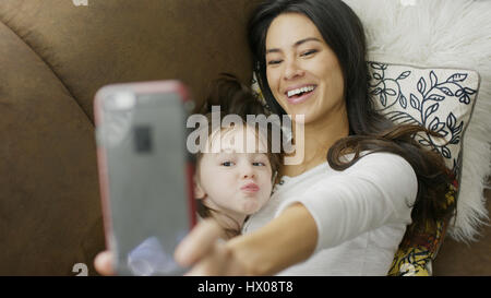 High angle selective focus view of playful mother and daughter posing and taking selfie with smartphone on sofa Stock Photo