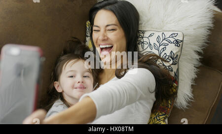 High angle selective focus view of playful mother and daughter posing and taking selfie with smartphone on sofa Stock Photo