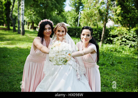 Bride posed on park with two cute brunette bridesmaids on pink dresses. Stock Photo