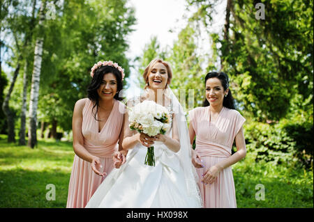 Bride posed on park with two cute brunette bridesmaids on pink dresses. Stock Photo