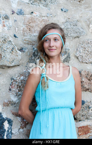 Young blonde woman in turqoise dress with braided hear and large blue earrings with  medieval stone walls behind Stock Photo