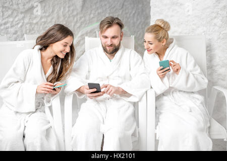 Three friends in bathrobes sitting with phones, applying salt therapy in the salt room Stock Photo