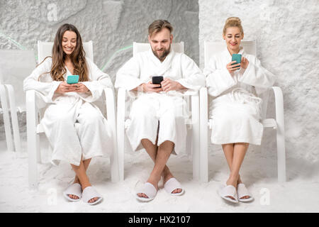 Three friends in bathrobes sitting with phones, applying salt therapy in the salt room Stock Photo