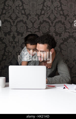 Little boy is climbing on his dad, trying to help him with his internet banking. They are sitting behind a laptop at home. Stock Photo