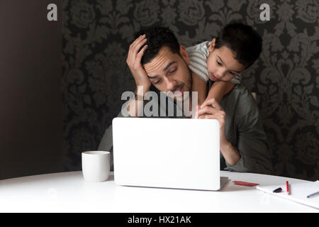 Little boy is climbing on his dad, trying to help him with his internet banking. They are sitting behind a laptop at home. Stock Photo