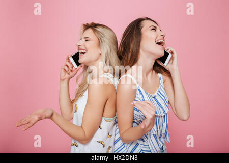 Two happy beautiful young women talking on mobile phones and laughing over pink background Stock Photo