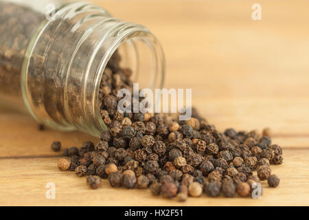 Black peppercorns is scattered on a wooden cutting board background from glass bottle. Stock Photo