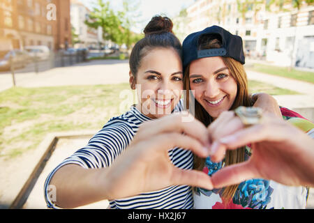 Two romantic young girlfriends making a heart gesture combining their hands as the pose arm in arm grinning at the camera Stock Photo
