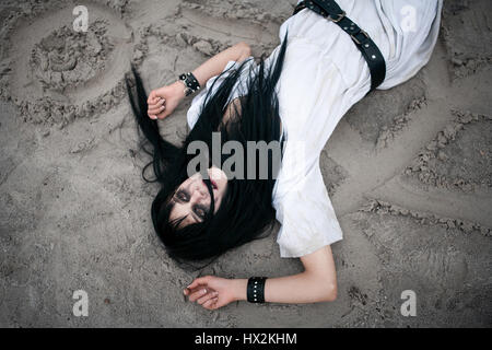 Crazy young woman lying on sand with painted symbols and looking up. Her eyes do not express anything. Close up. Stock Photo