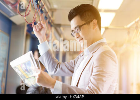 Asian Chinese businessman taking ride to work in morning, standing inside public transport and reading newspaper.