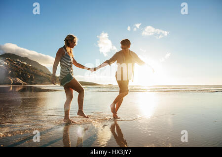 Full length shot of young couple holding hands walking on the beach and having fun. Young man and woman enjoying holidays on the sea shore. Stock Photo