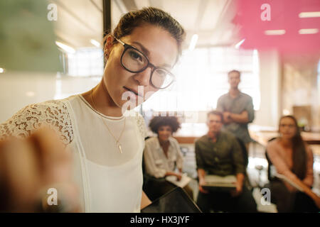 Businesswoman explaining new business ideas to colleagues while looking at sticky notes on glass wall. Female executive discussing business with team  Stock Photo